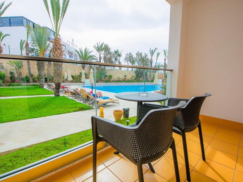 Balkon mit zwei Stühlen und Tisch, Blick auf grünen Garten und blauen Pool in sonniger Umgebung.