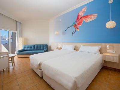 Camera d'hotel con due letti singoli, murale blu con uccello e area salotto con divano.