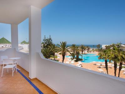 Balkon mit Blick auf einen Pool, Palmen und das Meer unter klarem Himmel.