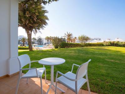 Zwei weiße Stühle und ein Tisch auf einer Terrasse mit Blick auf einen grünen Garten und den Pool im Hintergrund.