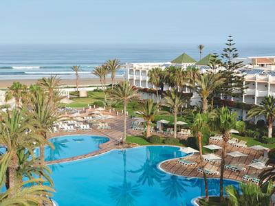 Blick auf einen Hotelpool mit Palmen und Meer im Hintergrund bei klarem Himmel.