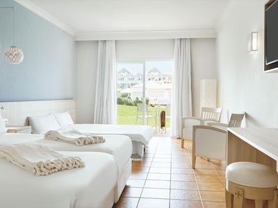 Helles Hotelzimmer mit zwei Einzelbetten, weißer Bettwäsche und Blick auf Terrasse und Garten.