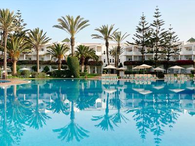 Grande piscina con palme e ombrelloni davanti a un edificio dell'hotel in una giornata serena.