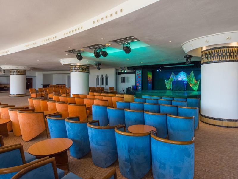 Sala conferenze con file di sedie arancioni e blu di fronte a un palco illuminato.