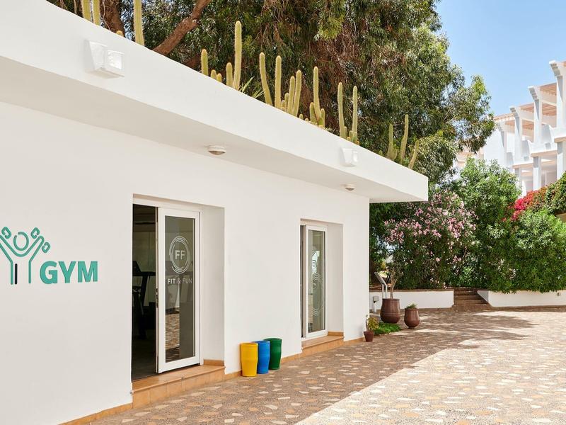 Edificio bianco della palestra con porta aperta e bidoni colorati all'esterno in una giornata di sole.