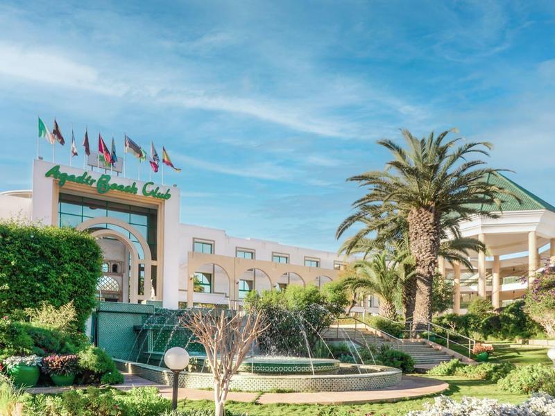 Bâtiment d'hôtel avec fontaine, palmiers et jardin vert sous ciel bleu