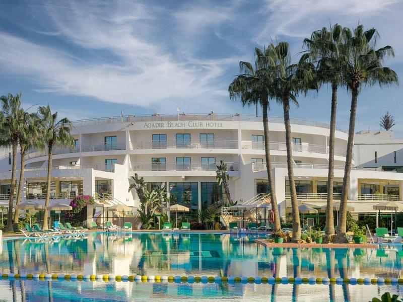 Grand bâtiment hôtelier blanc avec des palmiers et une piscine claire sous un ciel nuageux.