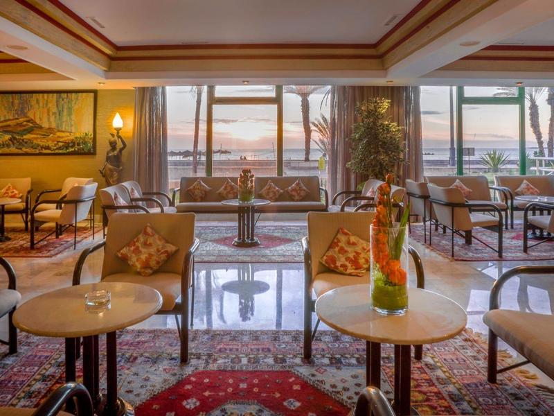 Salon d'hôtel élégant avec espaces de sièges, tapis et vue sur le coucher du soleil à travers de grandes fenêtres.