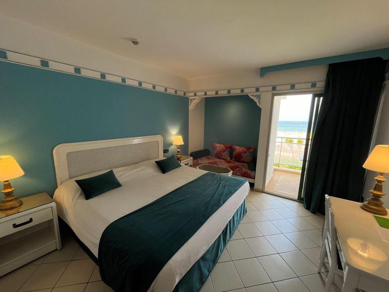 Chambre d'hôtel lumineuse avec lit double, coin salon, porte vers balcon avec vue sur mer.