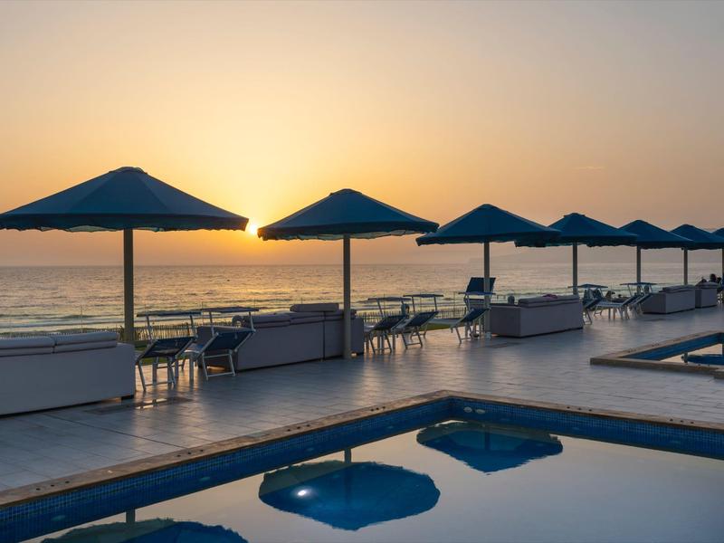 Pool mit blauer Wasserfläche, Sonnenschirme und Sitzbereich bei Sonnenuntergang am Meer.