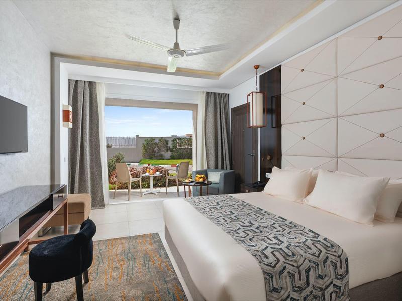 Modernes Hotelzimmer mit Doppelbett, Fernseher, Stuhl und Balkon mit Blick auf Garten und Meer.