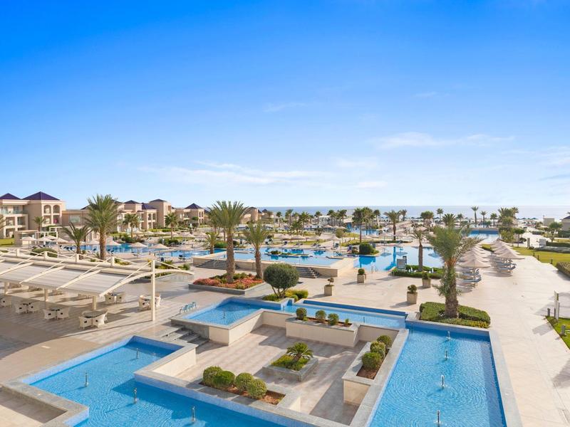 Hotelanlage mit Pools, Palmen und Wegen, blauer Himmel, moderne Architektur, offene Fläche.