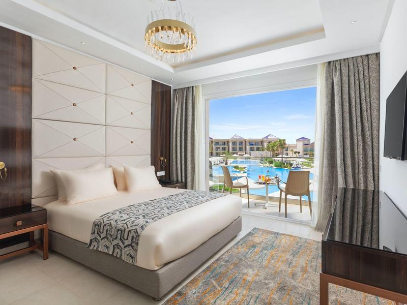 Helles Hotelzimmer mit großem Bett, Balkon und Blick auf einen Pool und Gebäude am Meer.