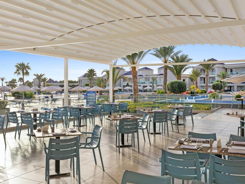Terrassenrestaurant mit blauen Stühlen und Tischen, Blick auf Pool und mediterrane Häuser.