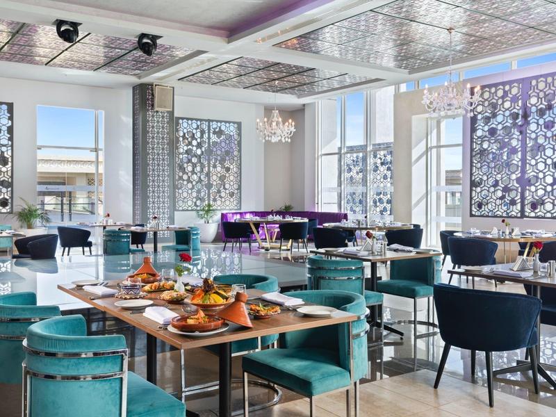 Modernes Restaurant mit großen Fenstern, blauen Stühlen, Holztischen und festlich gedeckten Platten.