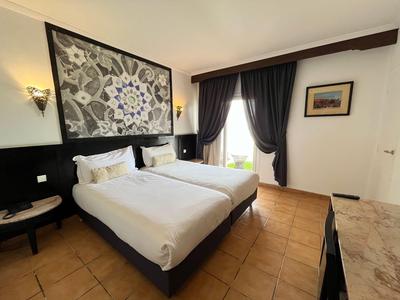 Habitación de hotel luminosa con dos camas individuales, tapiz decorativo y ventanas del suelo al techo con cortinas oscuras.