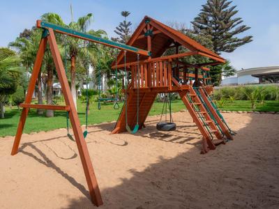 Parque infantil de madera con muro de escalada, columpio y tobogán en zona de arena de hotel.