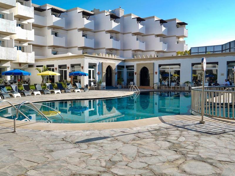 Moderner Hotelkomplex mit Außenpool, Liegestühlen und mehrstöckigen Balkonen bei Sonnenschein.