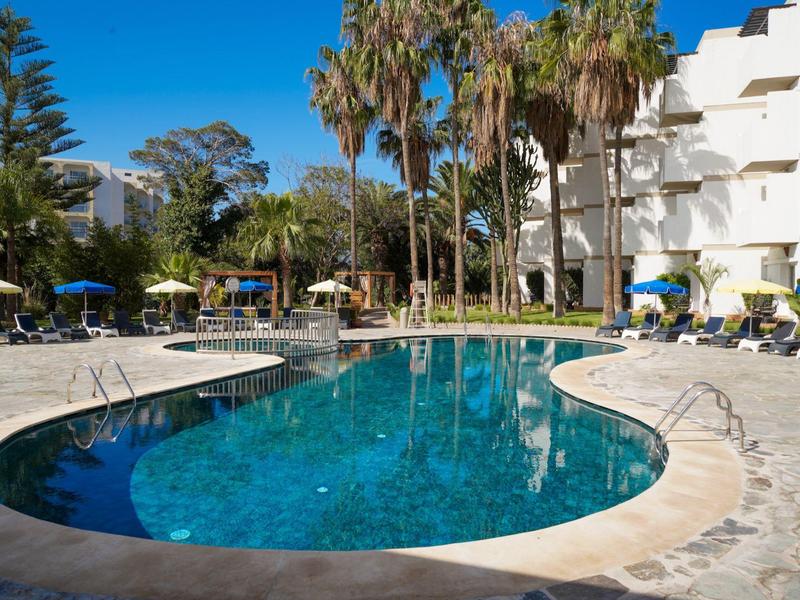 Runder Pool mit klarem Wasser, umgeben von Liegestühlen, Palmen und weißem Hotelgebäude bei blauem Himmel.