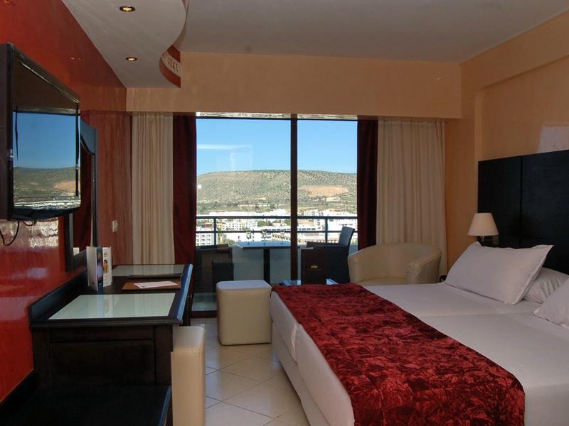 Moderní hotelový pokoj s manželskou postelí, červeným přehozem, balkonem a výhledem na krajinu.