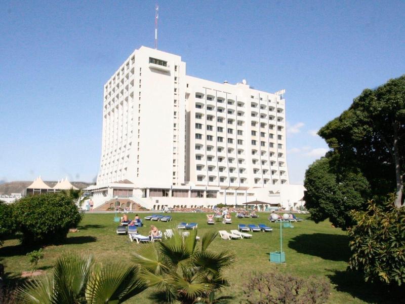 Vysoká bílá budova hotelu s velkými okny, zelenou zahradou a modrou oblohou.