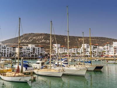 Segelboote im ruhigen Hafen mit weiß getünchten Gebäuden und Hügeln im Hintergrund.