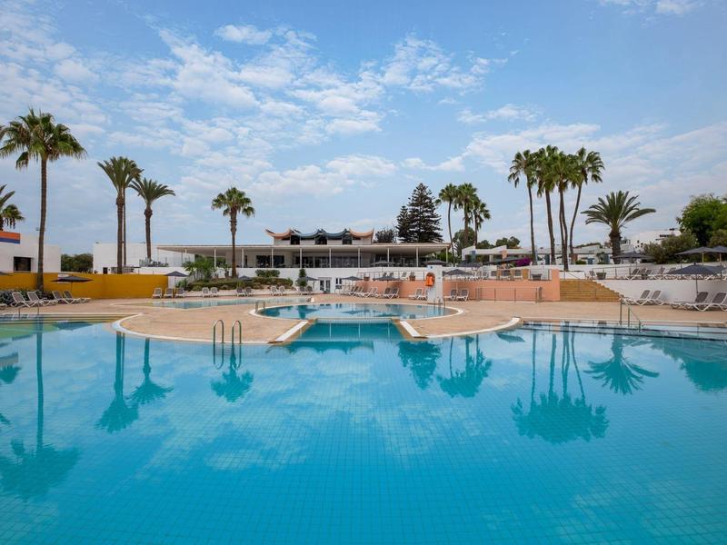 Großer Hotelpool mit Palmen und Sonnenterrasse unter blauem Himmel.