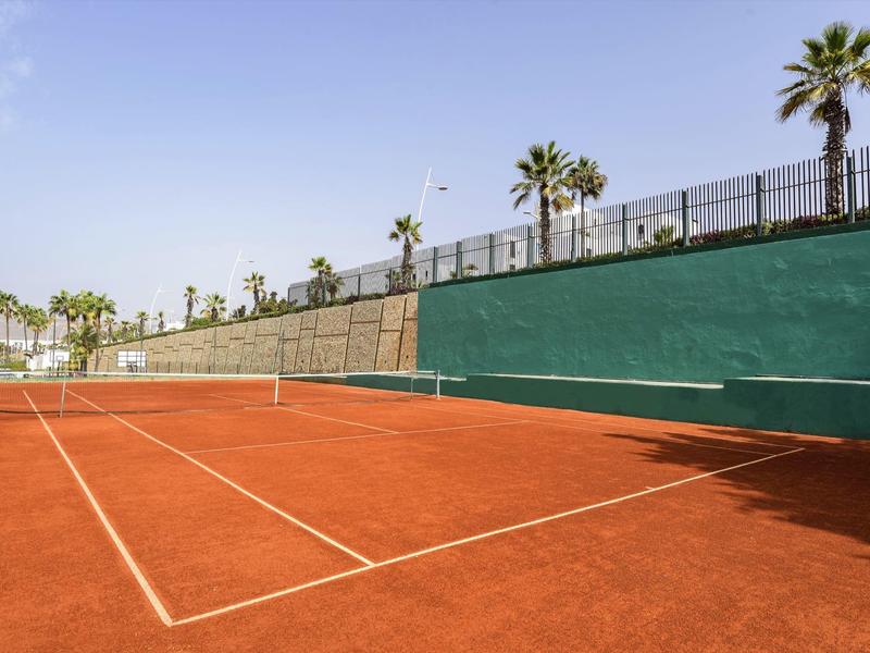 Roter Sandplatz-Tennisplatz mit Palmen und hellblauem Himmel im Freien.