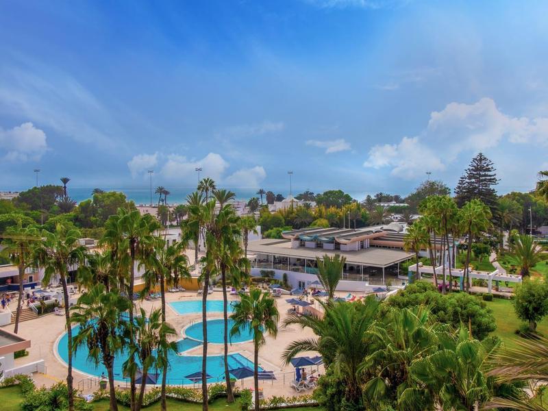 Hotelanlage mit mehreren Pools, Palmen und Sonnenschirmen unter blauem Himmel.
