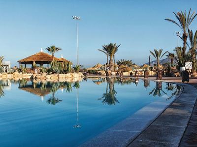 Groot zwembad met helder water, omringd door palmbomen en parasols onder een blauwe lucht.