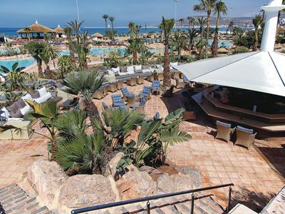 Vue sur une piscine d'hôtel tropicale avec des palmiers, parasols et mobilier de jardin au bord de la mer.