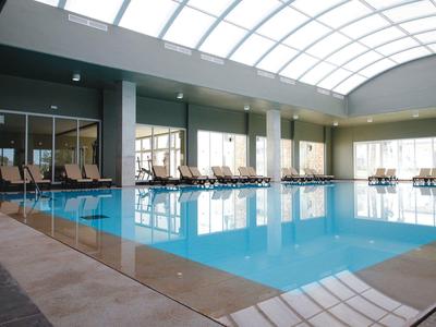 Grande piscine intérieure avec lumière naturelle, chaises longues et murs verts dans un hôtel.