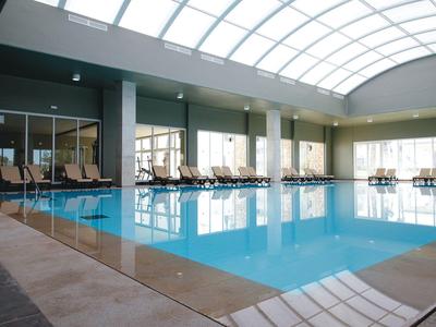 Grande piscine intérieure avec chaises longues et grandes fenêtres sous un toit vitré courbé.