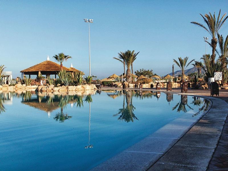 Grande piscine avec eau claire, entourée de palmiers et parasols sous un ciel bleu.