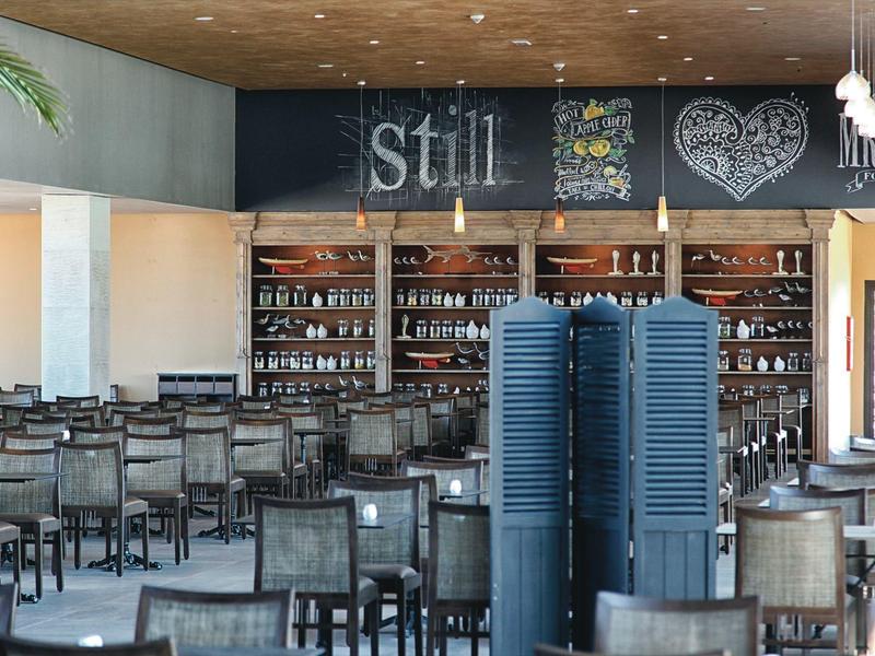 Salle à manger vide avec tables et chaises devant un bar avec un mur tableau noir marqué.