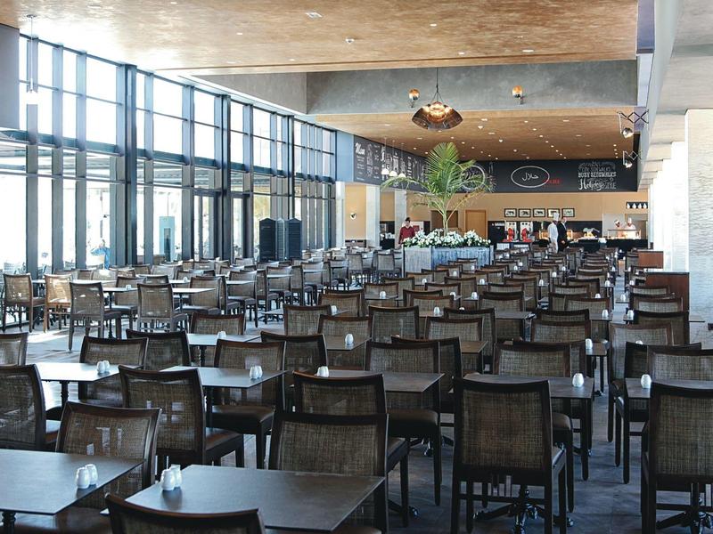 Grande salle à manger moderne avec de nombreuses chaises et tables, beaucoup de lumière naturelle par de grandes fenêtres.