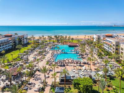 Großer Hotelkomplex mit Pool, Liegestühlen und Palmen direkt am weiten Sandstrand und blauem Meer.