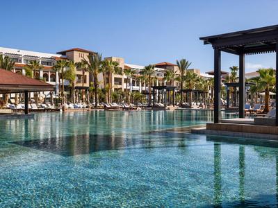 Großer Hotelpool mit Sonnenschirmen, Liegen und mehrstöckigem Gebäude im Hintergrund unter blauem Himmel