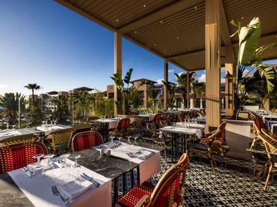 Terrassenrestaurant mit gedeckten Tischen und Blick auf Palmen bei sonnigem Wetter.
