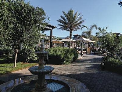 Ein schattiger Gartenweg mit einem steinernen Brunnen und Palmen, der zu einem Hotelbereich führt.