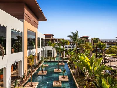 Moderner Hotelkomplex mit Swimmingpools, Palmen und üppiger Begrünung bei sonnigem Wetter.