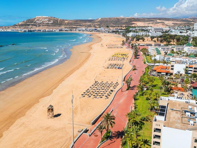 Lang zandstrand met ligbedden en omliggende hotels onder een blauwe lucht.