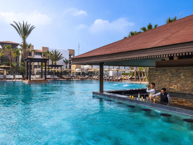 Groot hotelzwembad met swim-up bar en gasten, omgeven door palmbomen en parasols.