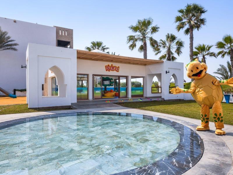 Hotel met zwembad, speeltuin en een vriendelijk verkleed personage in de zon.