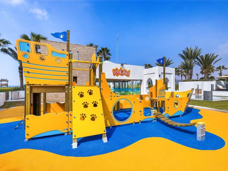 Kleurrijke speeltuin in de vorm van een schip met glijbaan en klimrekken onder een blauwe lucht.