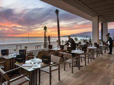 Offenes Restaurant mit Holzboden und Tischen, Blick auf Meer und bunten Himmel bei Sonnenuntergang.