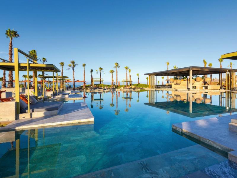 Großer Pool mit klar blauem Wasser, umgeben von Palmen und moderner, offener Architektur.
