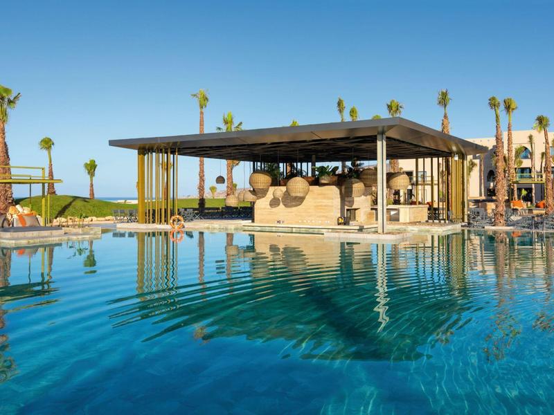 Großer Pool mit klarem Wasser, Sonnenschirmen und moderner Poolbar im Hintergrund bei blauem Himmel.