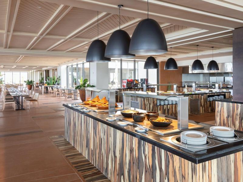 Großer moderner Speisesaal mit Buffet, Holzfußboden und schwarzen Hängeleuchten über Theke.