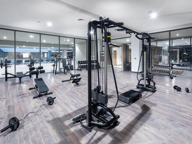 Moderner Fitnessraum mit Kraftmaschinen, Hanteln und großer Spiegelwand. Helle Holzoptik und weiße Wände.